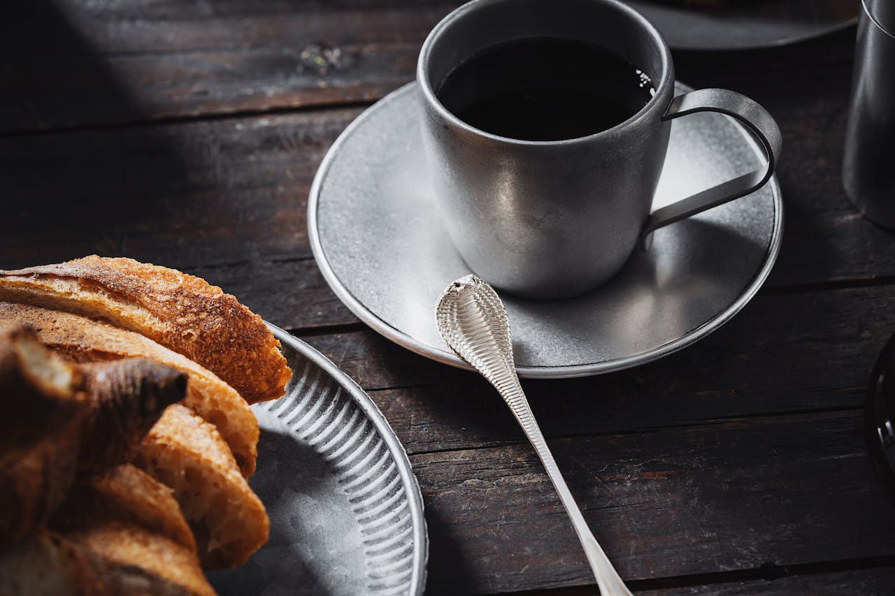 A cozy breakfast scene with silverware, bread, and a cup of coffee on a rustic table.