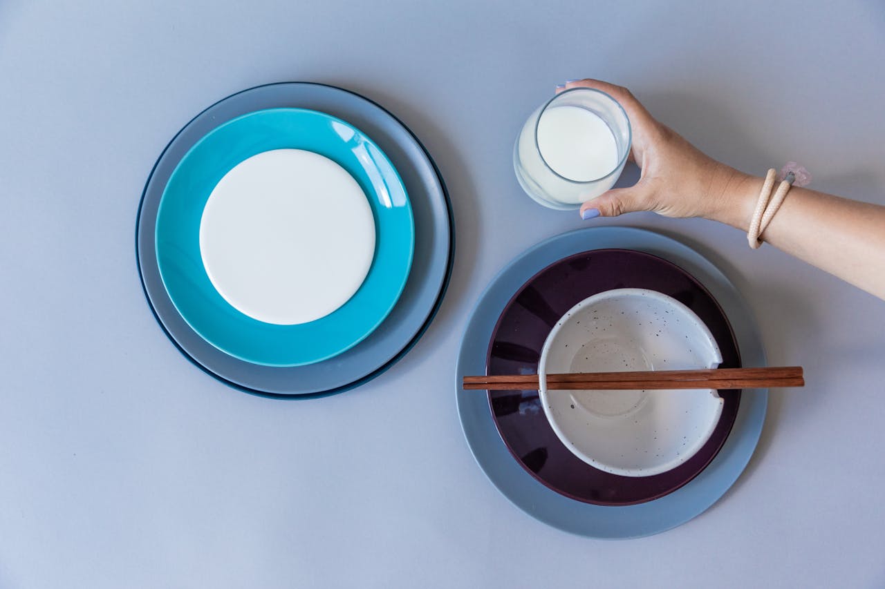 Top view of ceramic plates, a bowl with chopsticks, and a hand holding milk over a gray background.